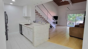 Eine moderne Küche mit einer zentralen Kochinsel, einem eingebauten Backofen und einem Edelstahlkühlschrank. Eine Treppe führt zu einem Wohnbereich mit einem großen Fenster und einem braunen Sofa.