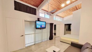 Ein geräumiger Wohnbereich mit hoher Decke, einem angebrachten Fernseher, einem Deckenventilator und einem großen Fenster, das für natürliches Licht sorgt.