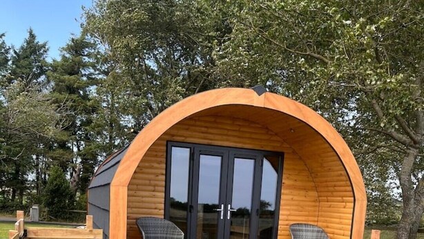 Una cabaña de madera con techo curvo, dos sillas y una mesa pequeña en el exterior.