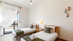 Een hotelkamer met twee bedden, een groot raam met gordijnen, een nachtkastje, een wandlamp en uitzicht op zee.