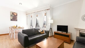 Een moderne woonkamer met een zwarte leren bank, een houten salontafel en een televisie op een houten dressoir. Er staat een eettafel met stoelen en een lamp.