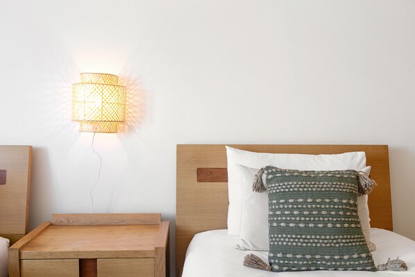 Una cama bien hecha con un cojín verde estampado y uno blanco, una mesita de noche de madera y una lámpara de pared.