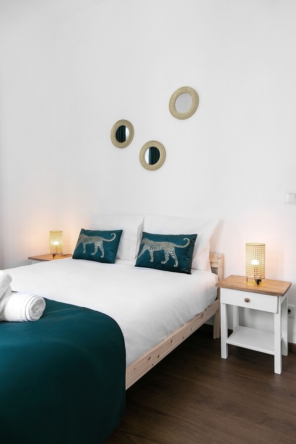Una cama bien hecha con ropa de cama blanca y almohadas turquesa, dos mesitas de noche con lámparas, y dos espejos de pared.