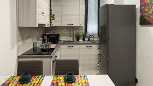 A modern kitchen with light wood cabinets, a stainless steel refrigerator, a coffee maker, and a dining area with a table and chairs.