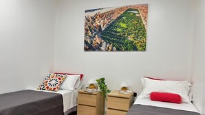 A hotel room with two single beds, a red rug, a nightstand with a lamp, a small plant, and a painting of a cityscape.