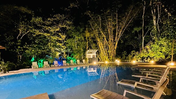A nighttime swimming pool with lounge chairs and a small building, surrounded by trees and greenery. - Grand Park Manuel Antonio (Quepos)