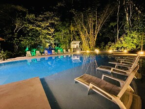 Una piscina nocturna con sillones de descanso y un pequeño edificio, rodeada de árboles y vegetación.