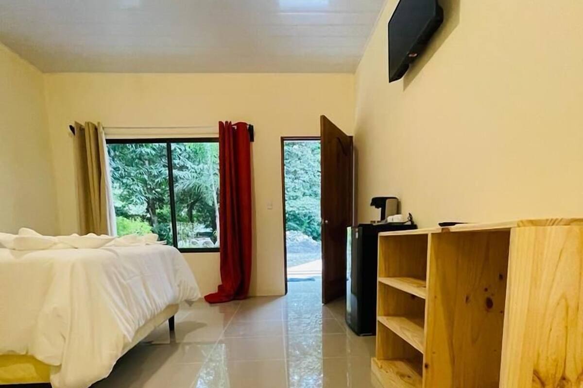 A hotel room with a bed, a TV mounted on the wall, a wooden shelf unit, and a window with curtains.