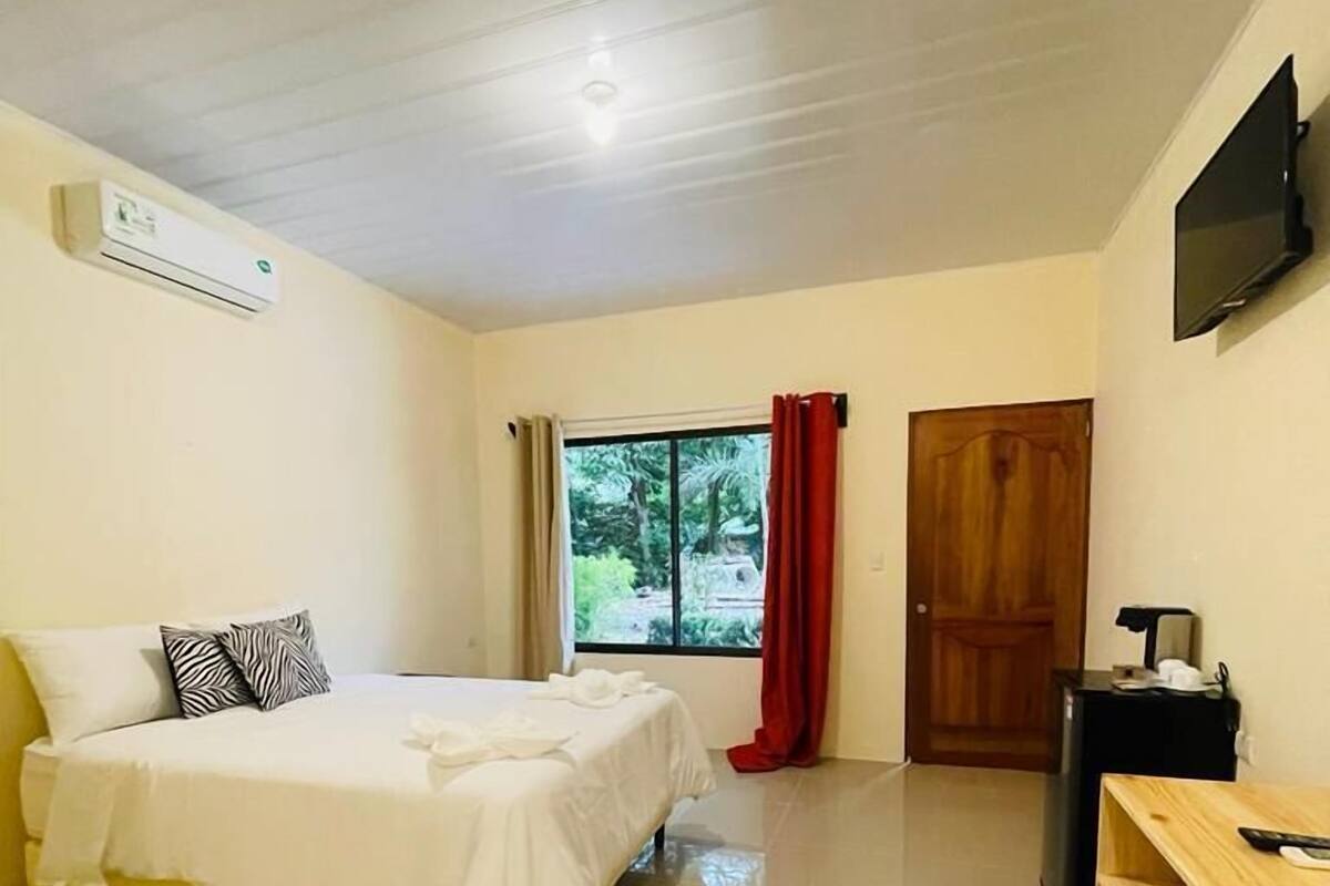 A bedroom with a bed, a window with curtains, an air conditioning unit, a TV, and a wooden side table.