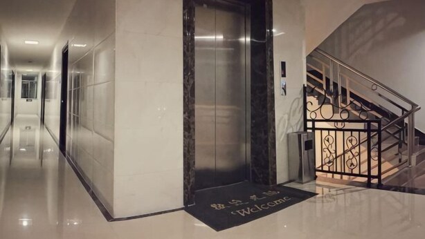 Een hotel lobby met een marmeren vloer, een zwarte lift, een trap met een metalen leuning, en een welkomstmat.