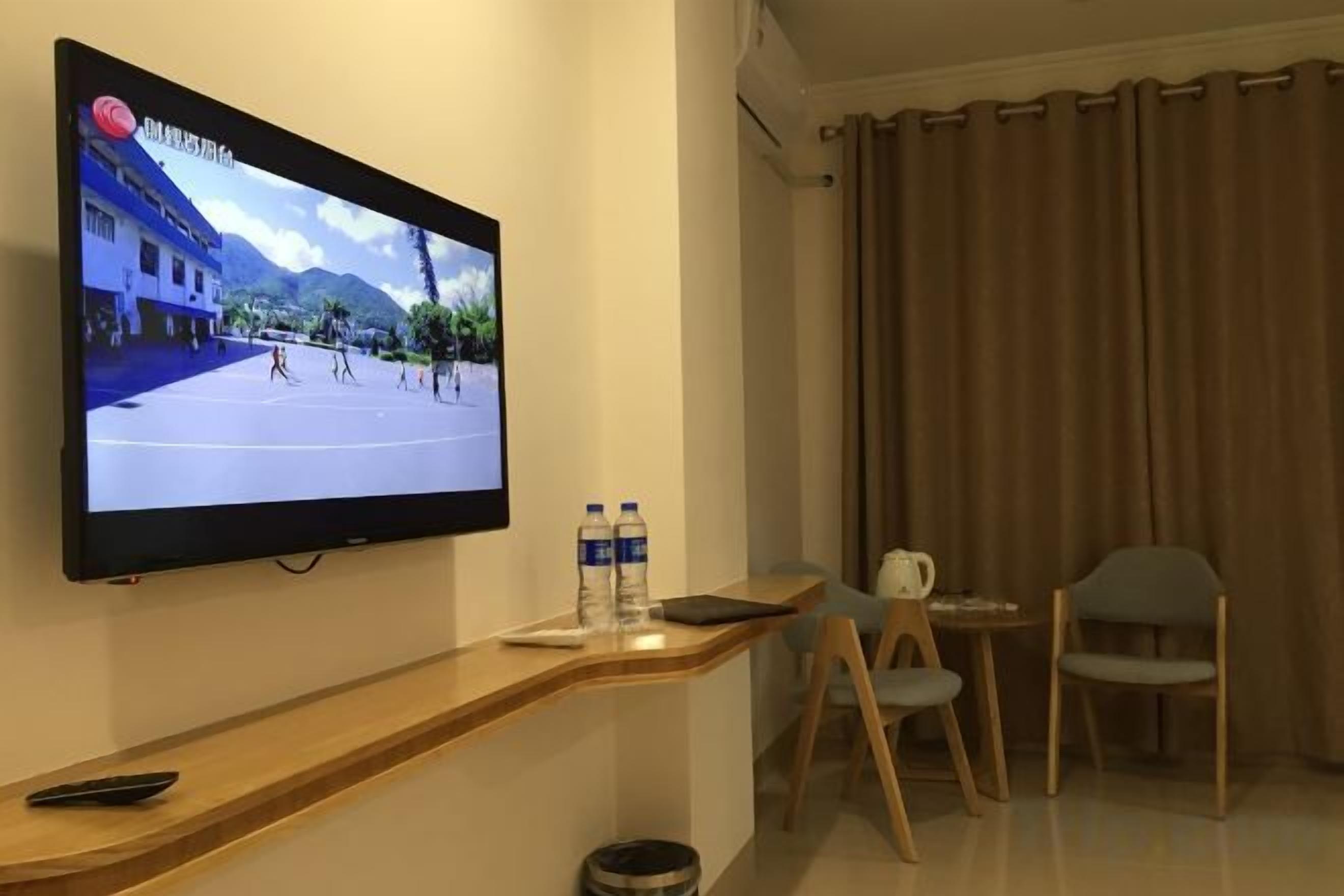 A hotel room with a flat-screen TV mounted on the wall, a wooden console table with a remote control, two chairs, and a small table with a water bottle and a tablet.