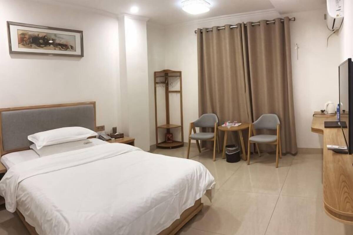 A hotel room with a bed, two chairs, a desk, a television, and a painting on the wall.