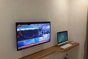 Ett rum med en stor tv-skärm med platt skärm som hänger på väggen och visar finansiell information, en mindre bildskärm på ett skrivbord i trä samt ett tangentbord.