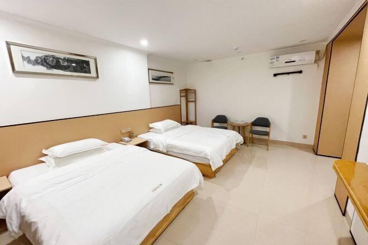 A hotel room with two single beds, a wooden headboard, a small table with a chair, and a wall-mounted air conditioning unit.