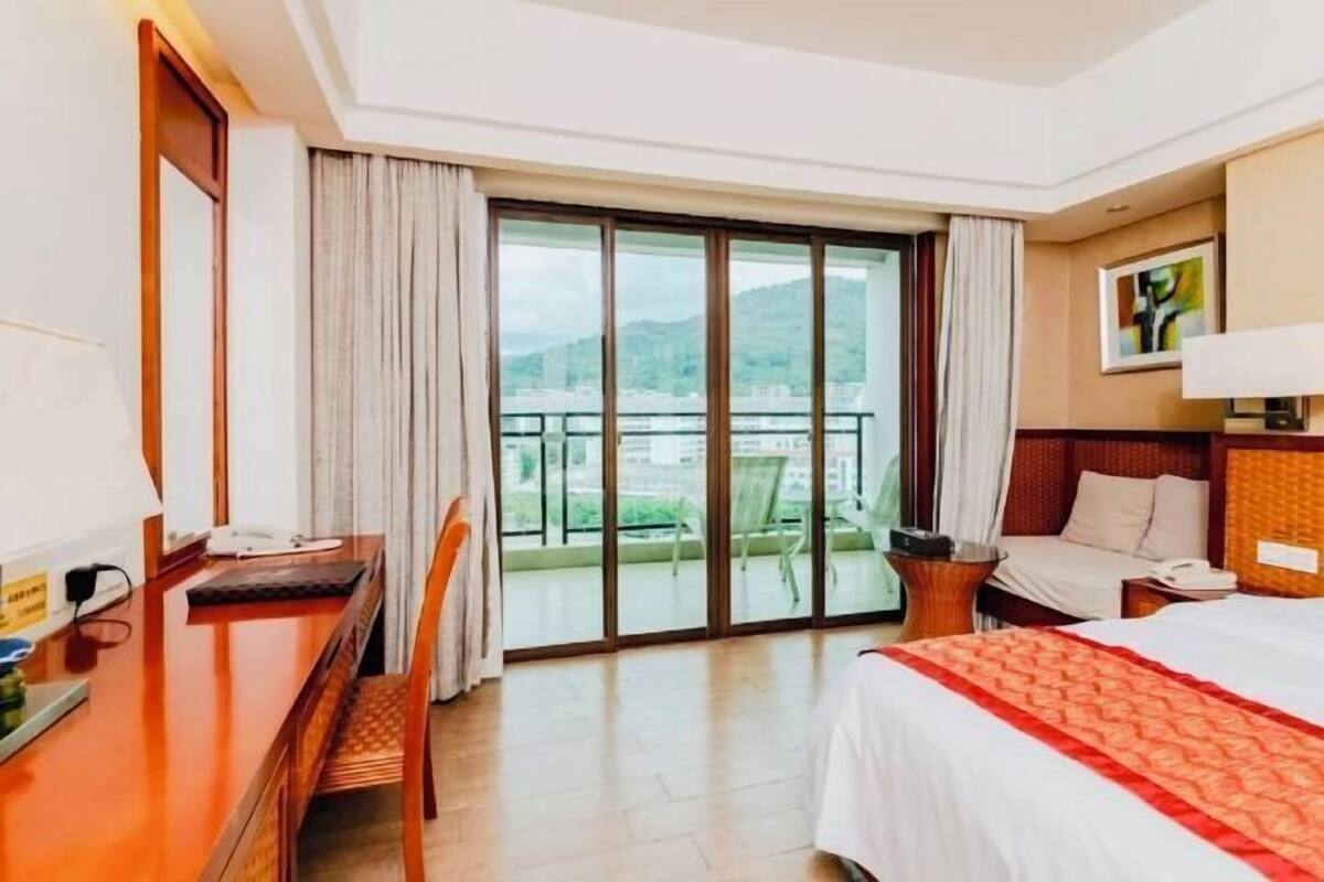 A hotel room with a bed, a desk, a chair, a large window with a view of mountains, and a balcony.