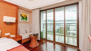 Una habitación de hotel con cama, mesita de noche, una pequeña mesa redonda, una silla, balcón con vistas a la ciudad y un cuadro en la pared.