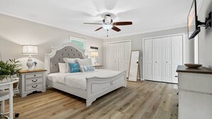 A bedroom with a white bed, a ceiling fan, a nightstand with a lamp, a dresser, and a TV mounted on the wall.