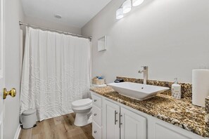 Ein Badezimmer mit Toilette, Waschbecken mit Granitplatte, einer Duschvorhang und einem kleinen Mülleimer.