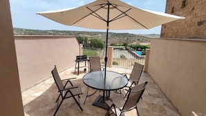 Une table ronde, quatre chaises et un grand parasol sur un balcon offrant une vue sur une piscine et des collines.