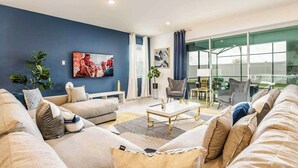 A modern living room with a blue accent wall, a large flat-screen TV, a dining area with a glass table, and a view through large windows. - Huge 9BR Vacation Villa in Orlando | Near Disney (Kissimmee)