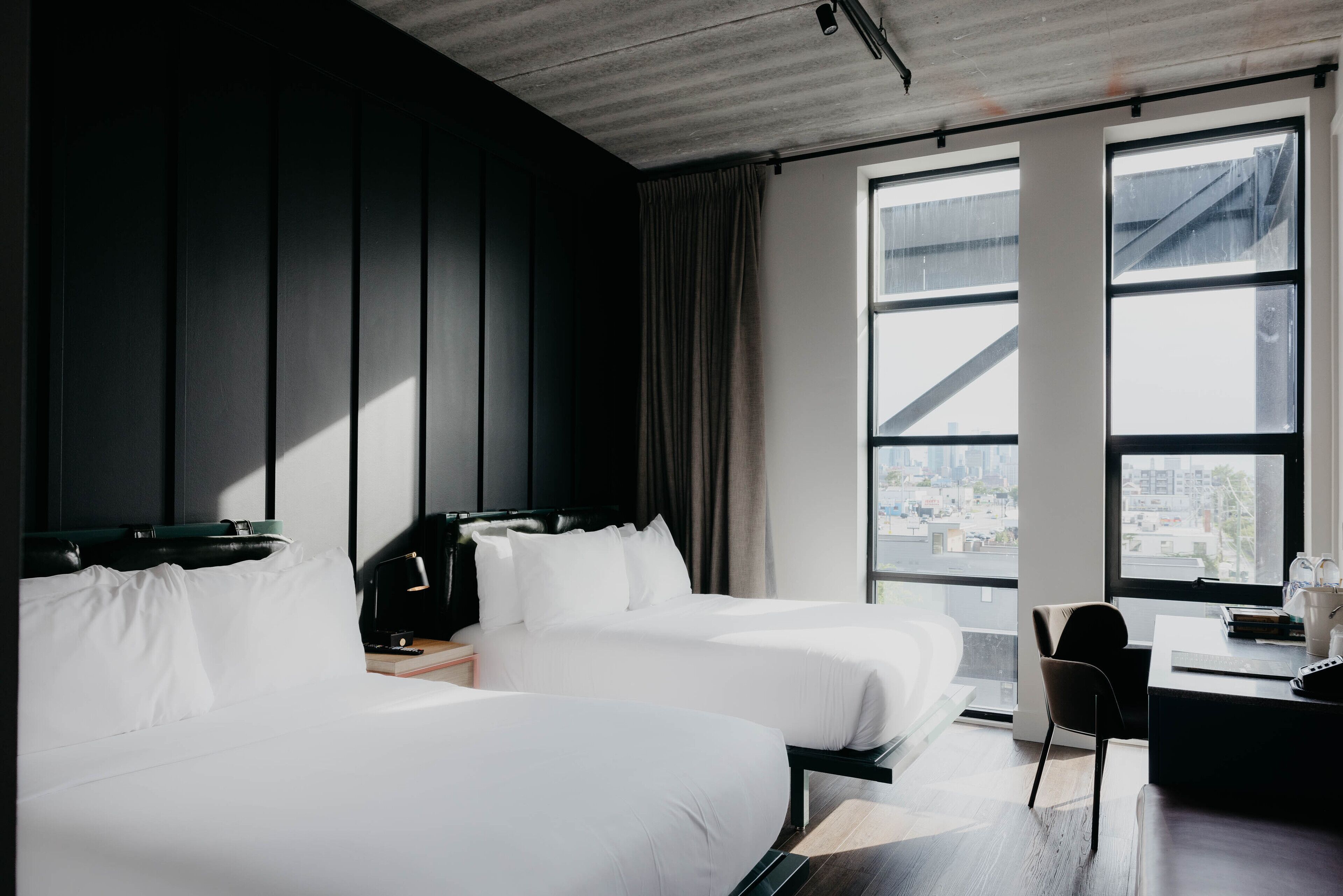 Una habitación de hotel moderna con dos camas, un escritorio y ventanas grandes que ofrecen vistas a la ciudad.
