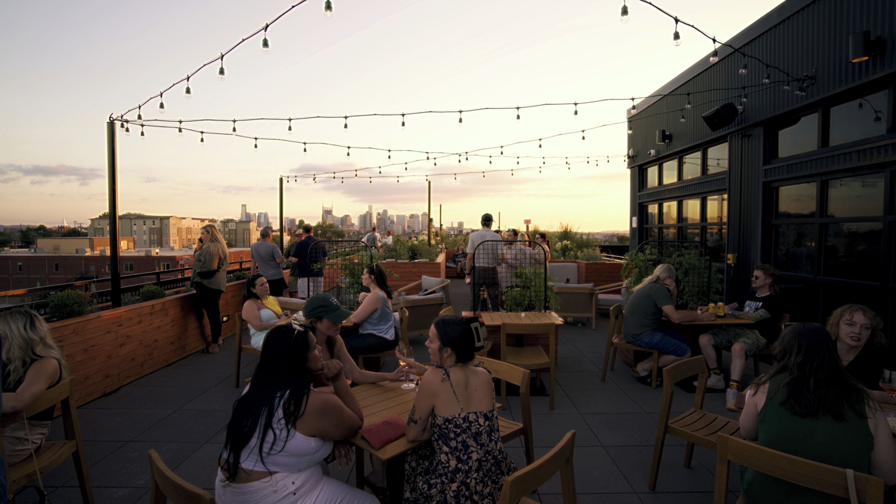 An outdoor seating area with tables and chairs, string lights overhead, and a city skyline in the background.
