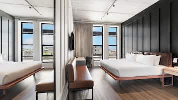 Una habitación de hotel moderna con dos camas, un banco, un escritorio y grandes ventanales con vistas a la ciudad.