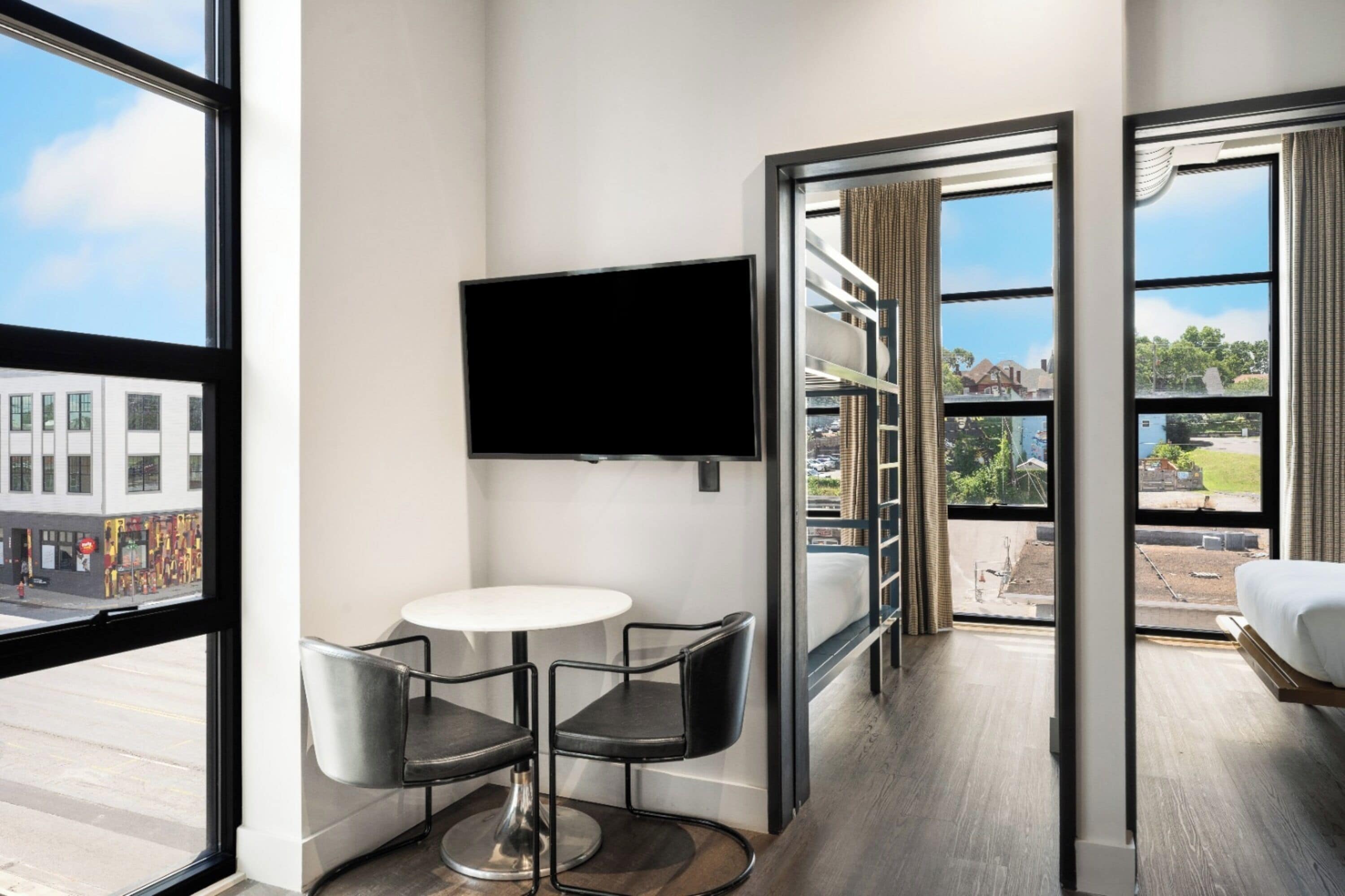 A hotel room with a large window, a flat-screen TV mounted on the wall, a small round table, two chairs, and a bed with a view of the outside.
