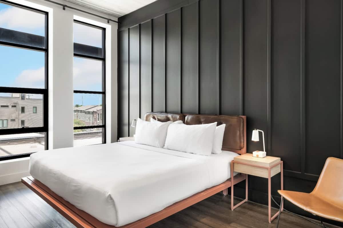 A modern bedroom with a large bed, a nightstand with a lamp, and a view of buildings through large windows.