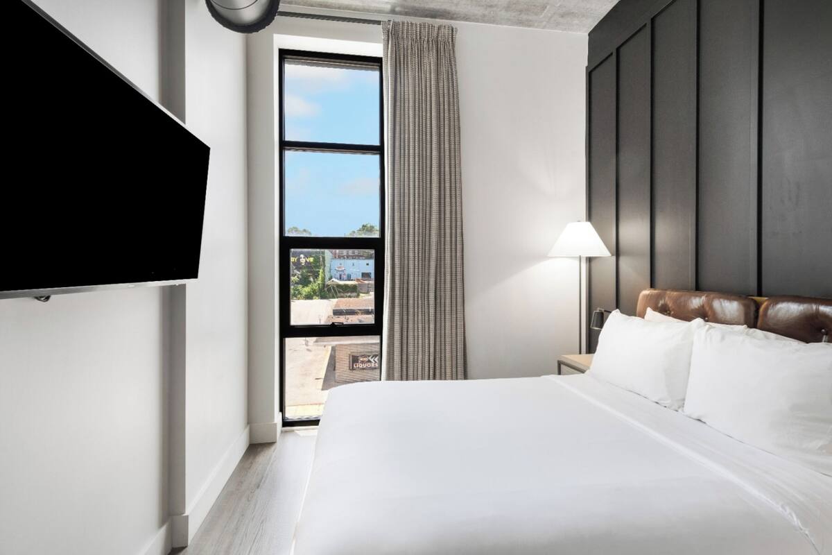 A hotel room with a large bed, a TV mounted on the wall, a window with curtains, and a view of the outside.