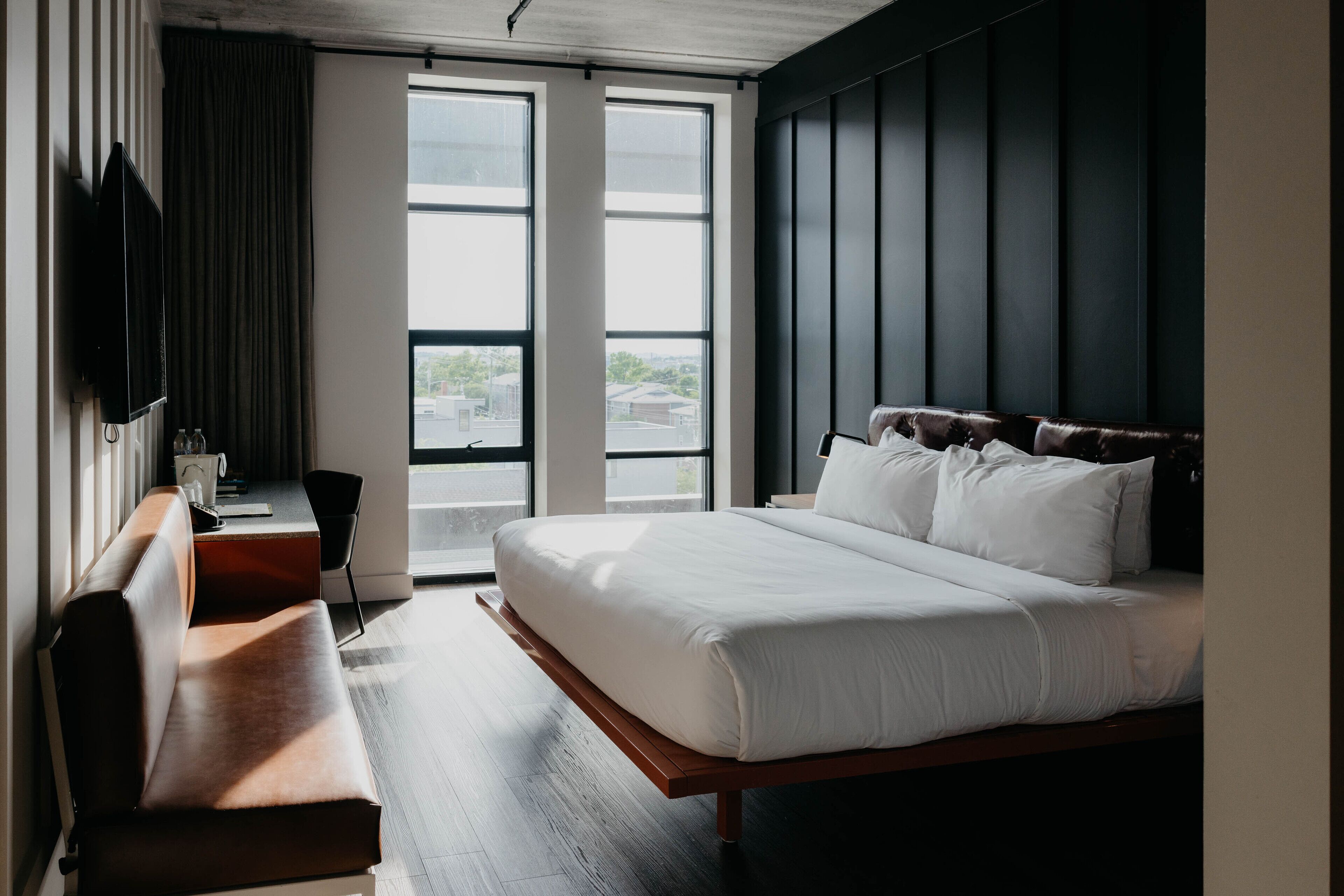 A modern hotel room with a large bed, a wooden bench, a TV, and a view of the city through large windows.