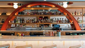 A bar with a curved wooden structure, shelves of bottles, glasses, and a bar counter. There are three bar stools in front of the counter.