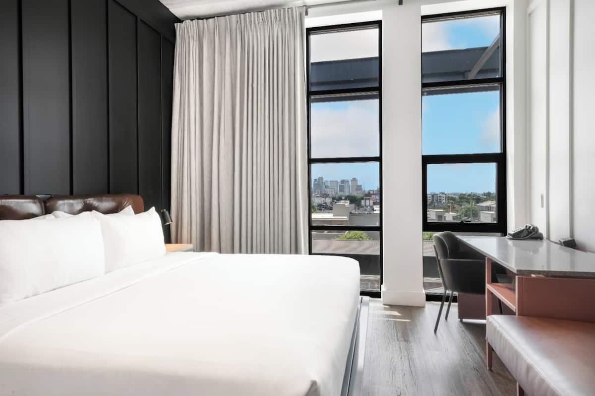A modern hotel room with a large bed, a desk, and a view of the city through large windows.