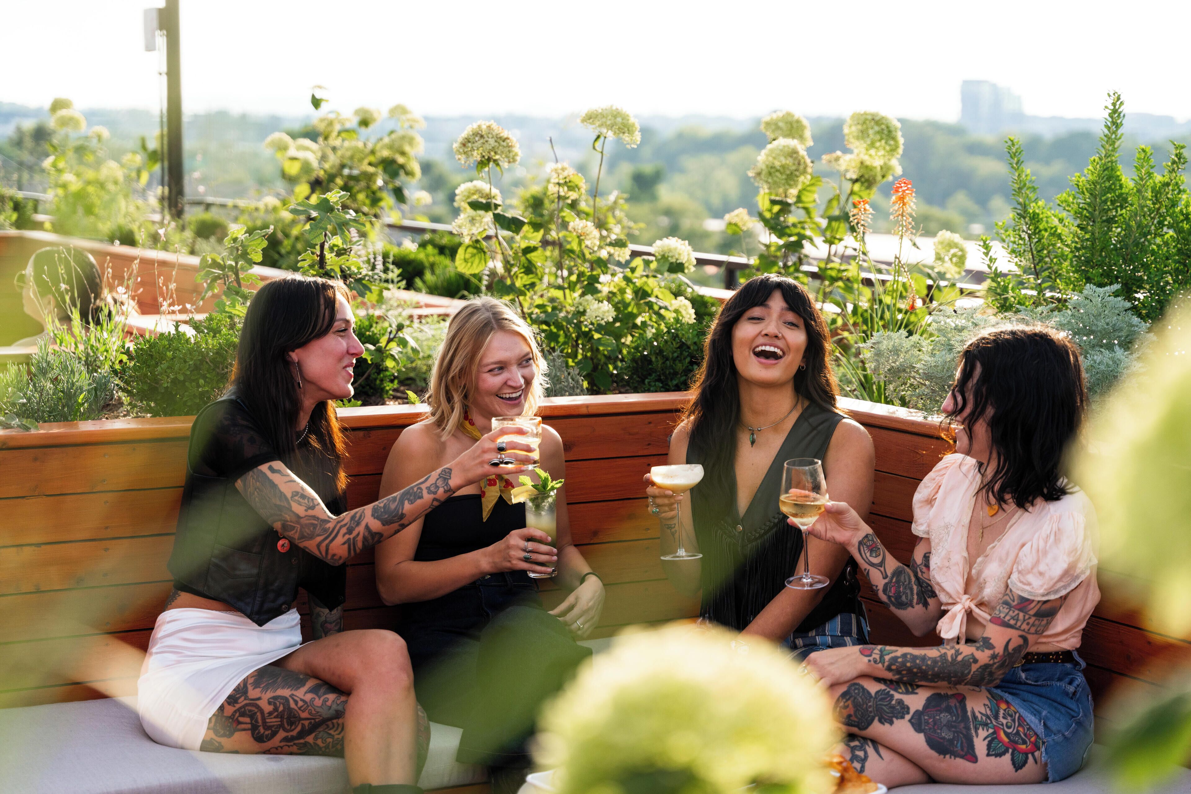 A group of people sitting on a wooden bench outdoors with a view of greenery and buildings in the background.