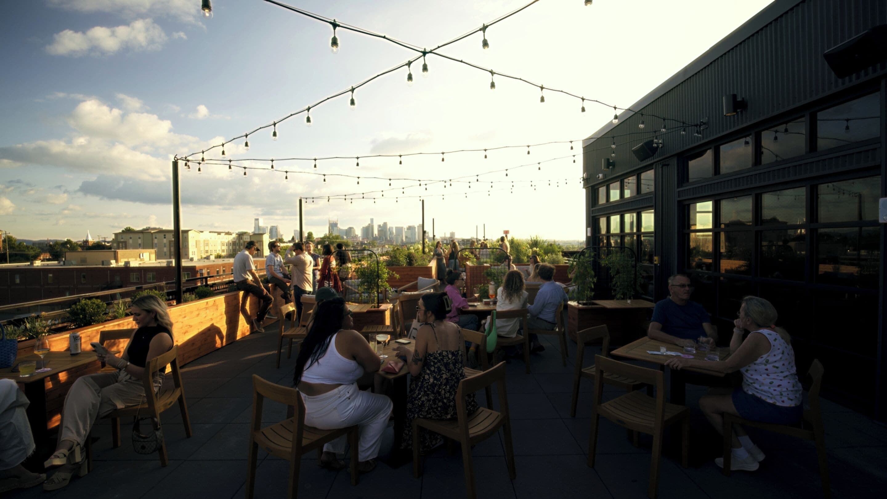 An outdoor seating area with tables and chairs, string lights overhead, and a city skyline in the background.