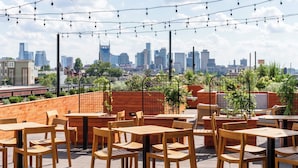 An outdoor seating area with tables and chairs, string lights, and a city skyline in the background.