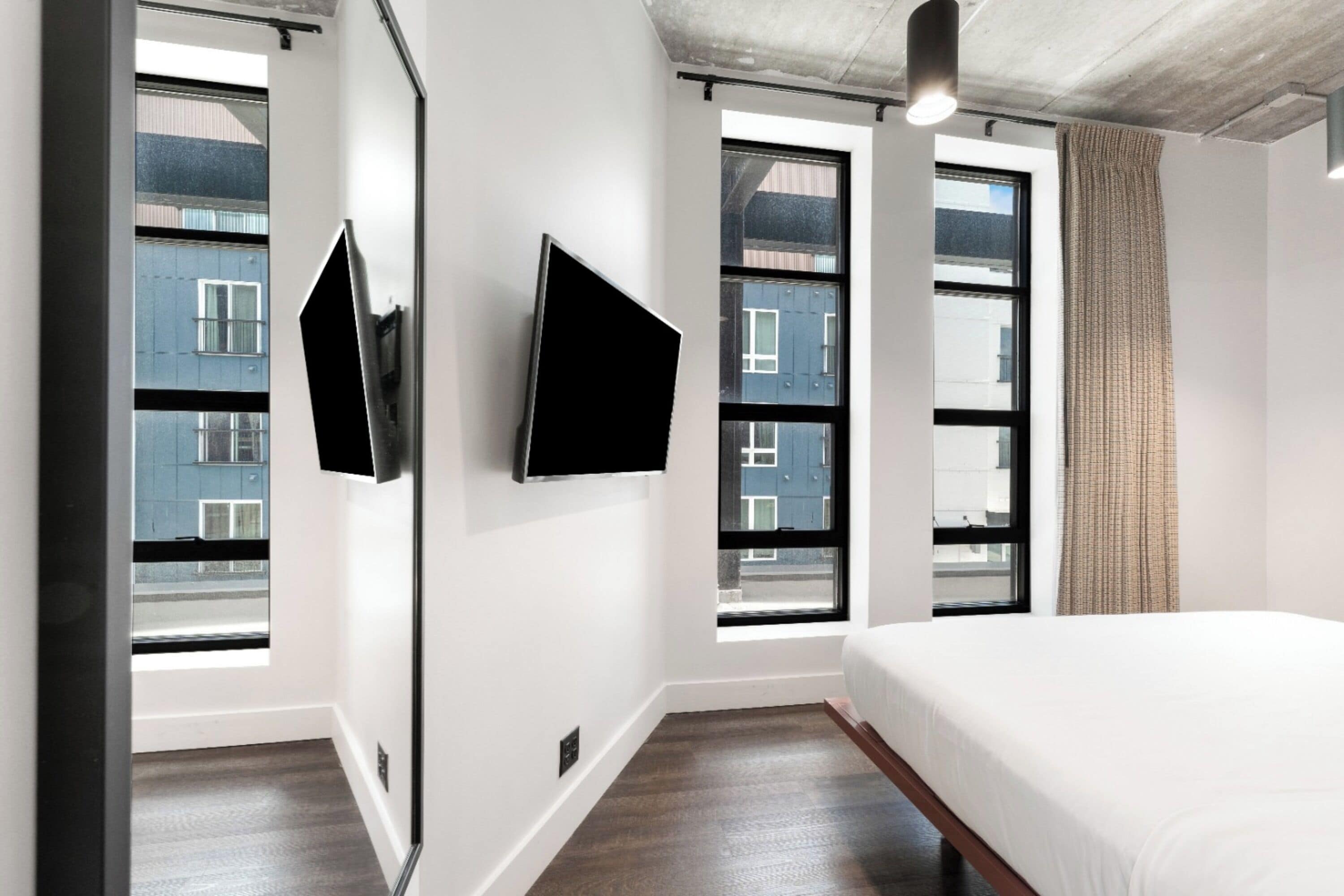 Ein modernes Hotelzimmer mit einem großen Bett, zwei Fenstern und einem an der Wand befestigten Flachbildfernseher.