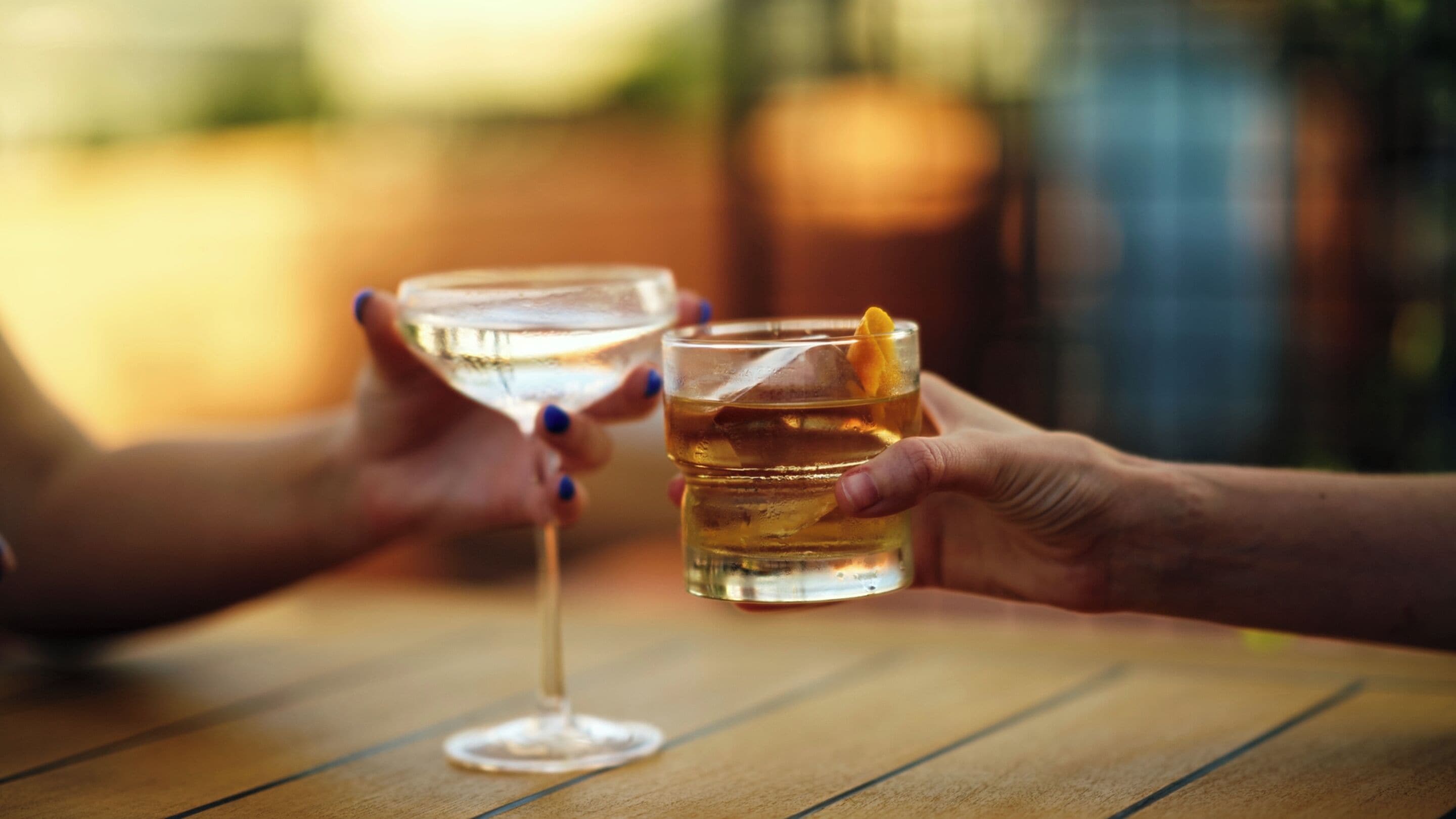 Two people holding glasses of a clear liquid, possibly a drink, on a wooden table outdoors.
