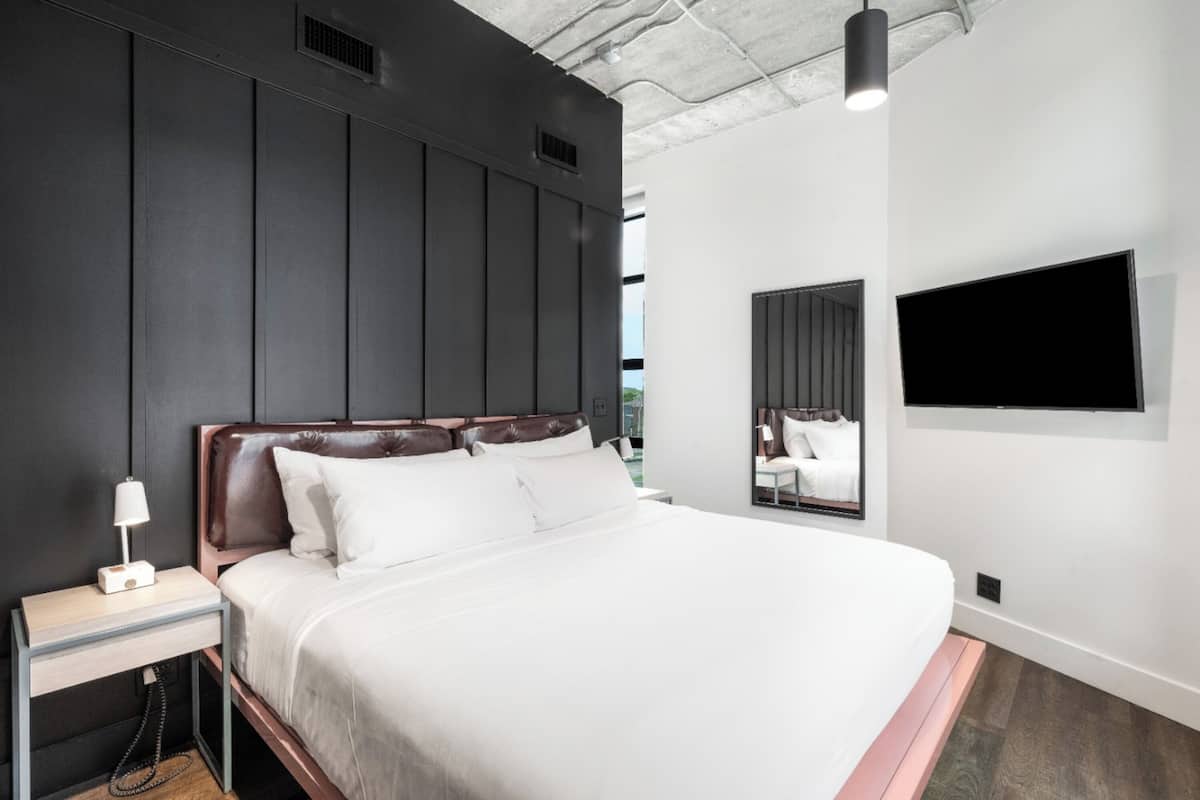 A modern hotel room with a large bed, a bedside table with a lamp, a flat-screen TV mounted on the wall, and a large mirror.