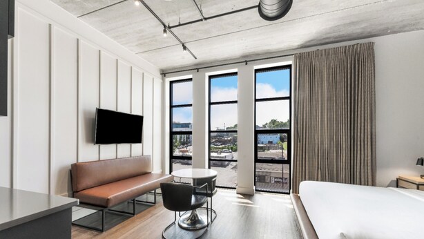 A modern hotel room with a large bed, a sofa, a dining table, and a TV mounted on the wall.