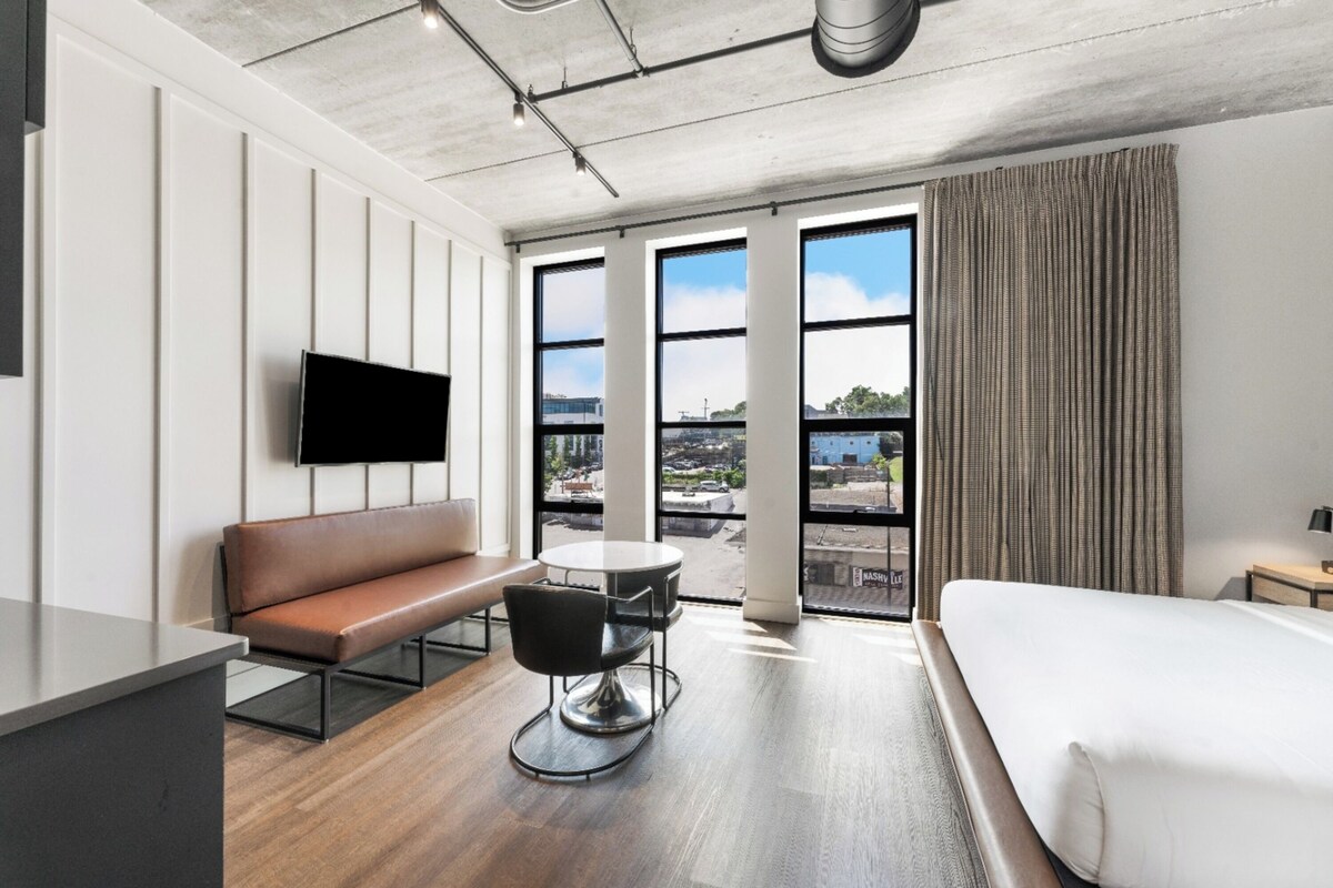 A modern hotel room with a large bed, a sofa, a dining table, and a TV mounted on the wall.