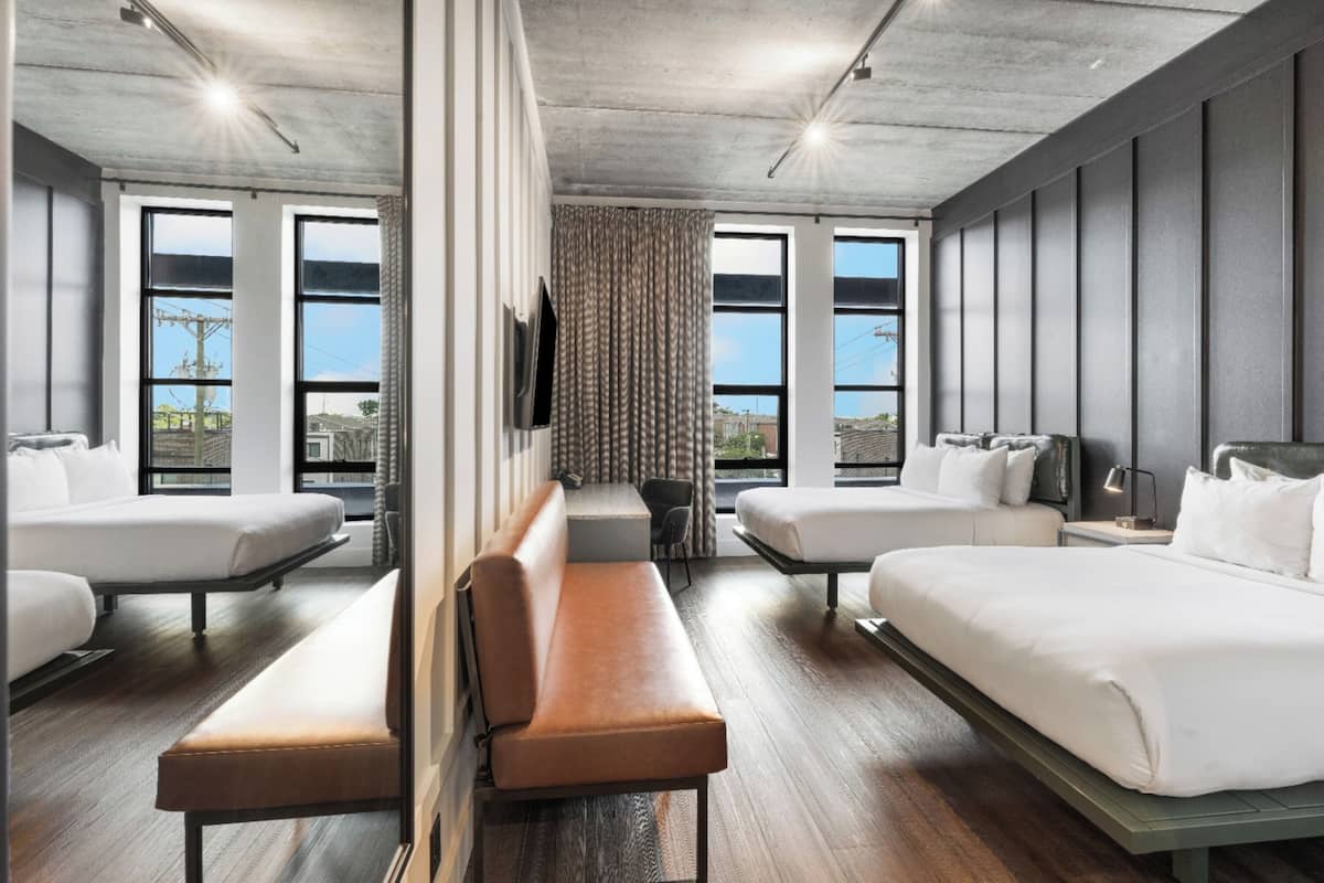 A modern hotel room with two beds, a bench, a desk, and large windows.