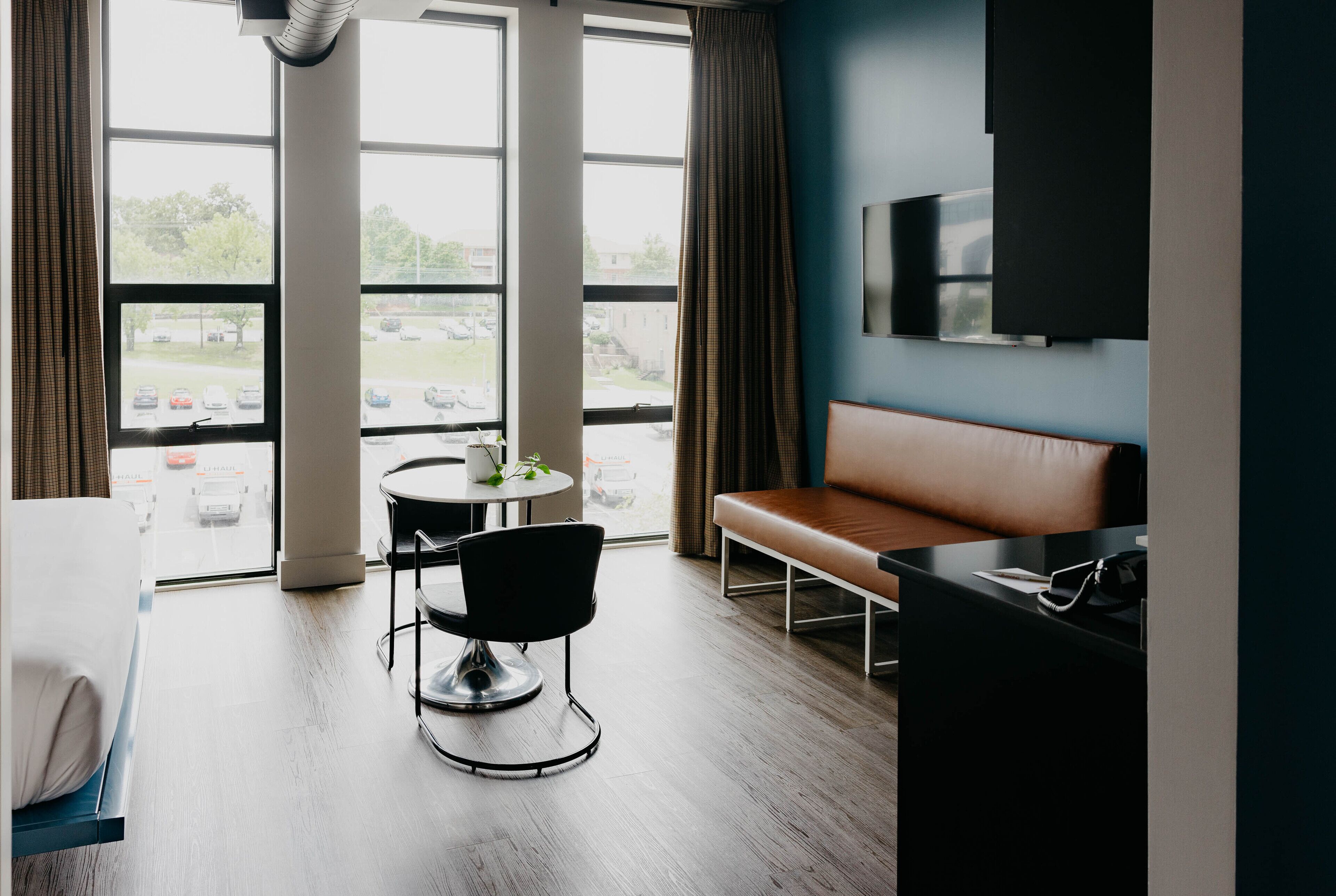 Ein Hotelzimmer mit großem Fenster, einer Couch, einem Esstisch mit Stuhl und einem an der Wand befestigten Fernseher.