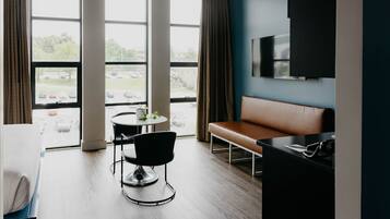 Ein Hotelzimmer mit großem Fenster, einer Couch, einem Esstisch mit Stuhl und einem an der Wand befestigten Fernseher.
