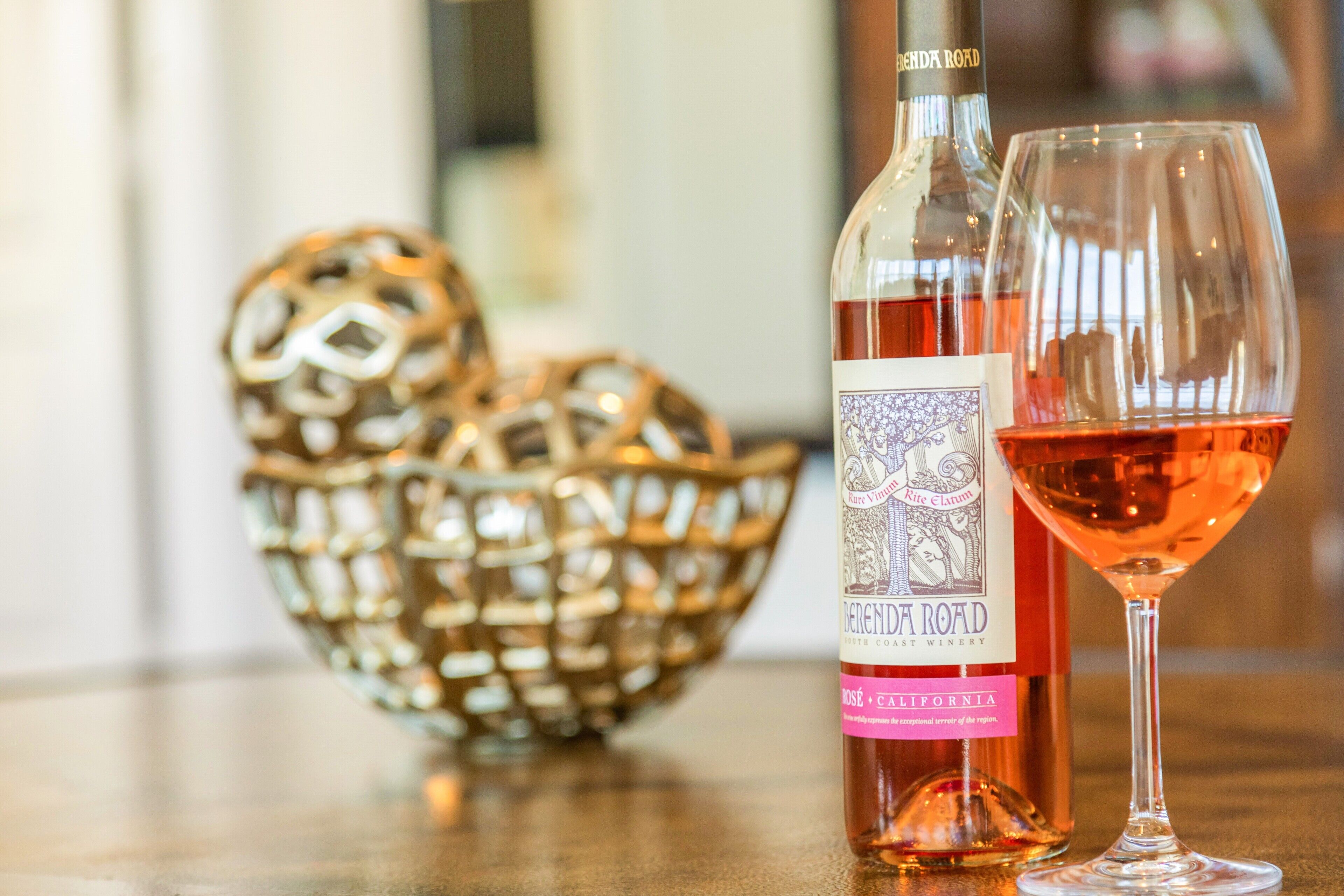 A bottle of wine and a wine glass on a table with a decorative bowl in the background.