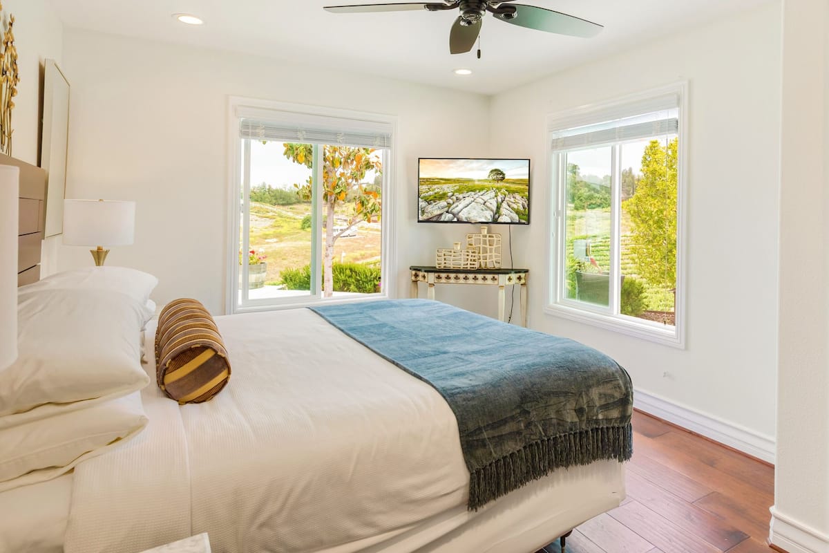 A bedroom with a large bed, a ceiling fan, two windows with a view of greenery, and a nightstand with a lamp.