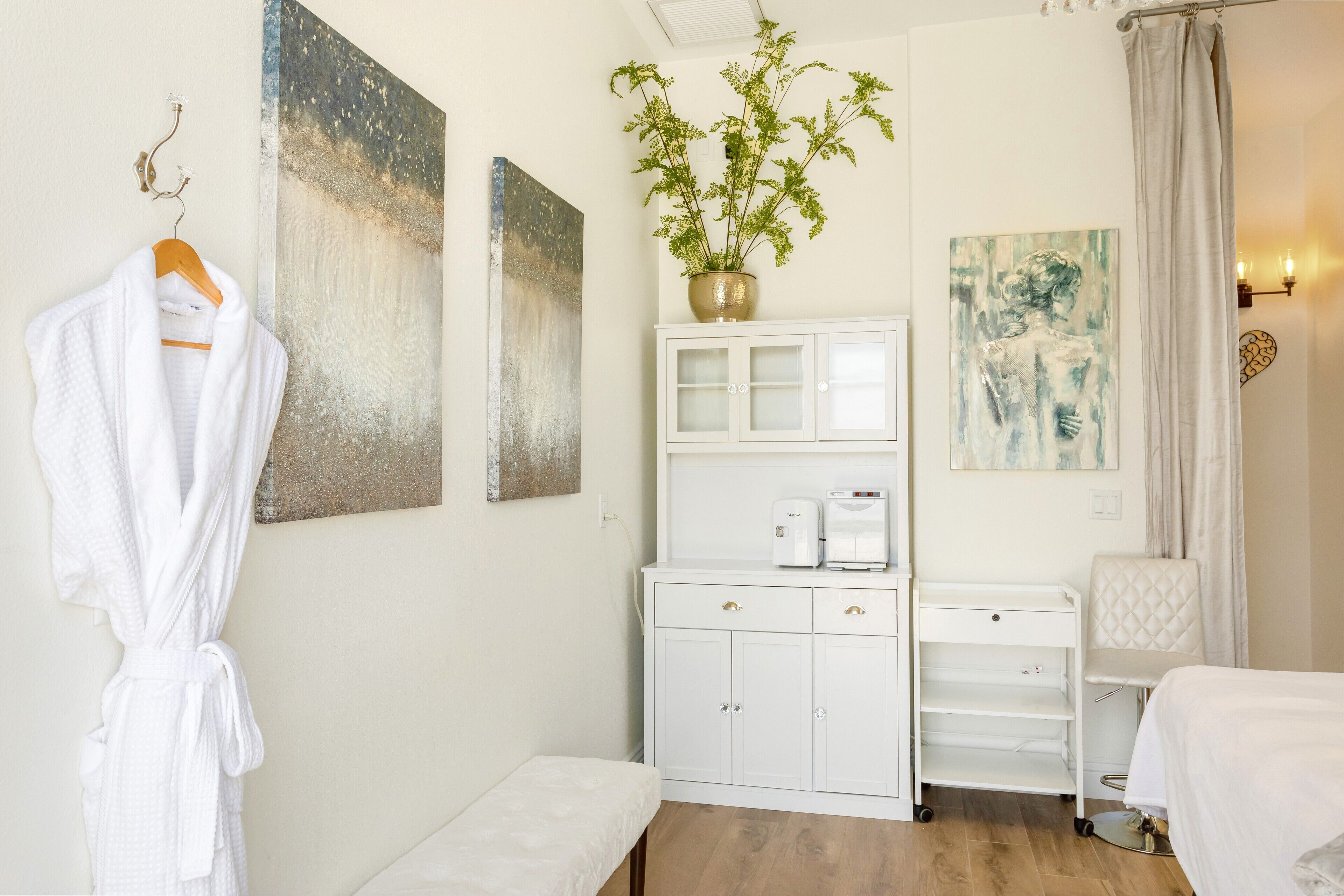 Een hotelkamer met een witte badjas hangend aan een haak, een wit kastje met lades, een klein tafeltje, een witte stoel en een bed.