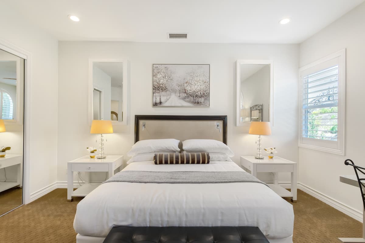 A bedroom with a large bed, two bedside tables with lamps, a headboard, a mirror, and a window with blinds.