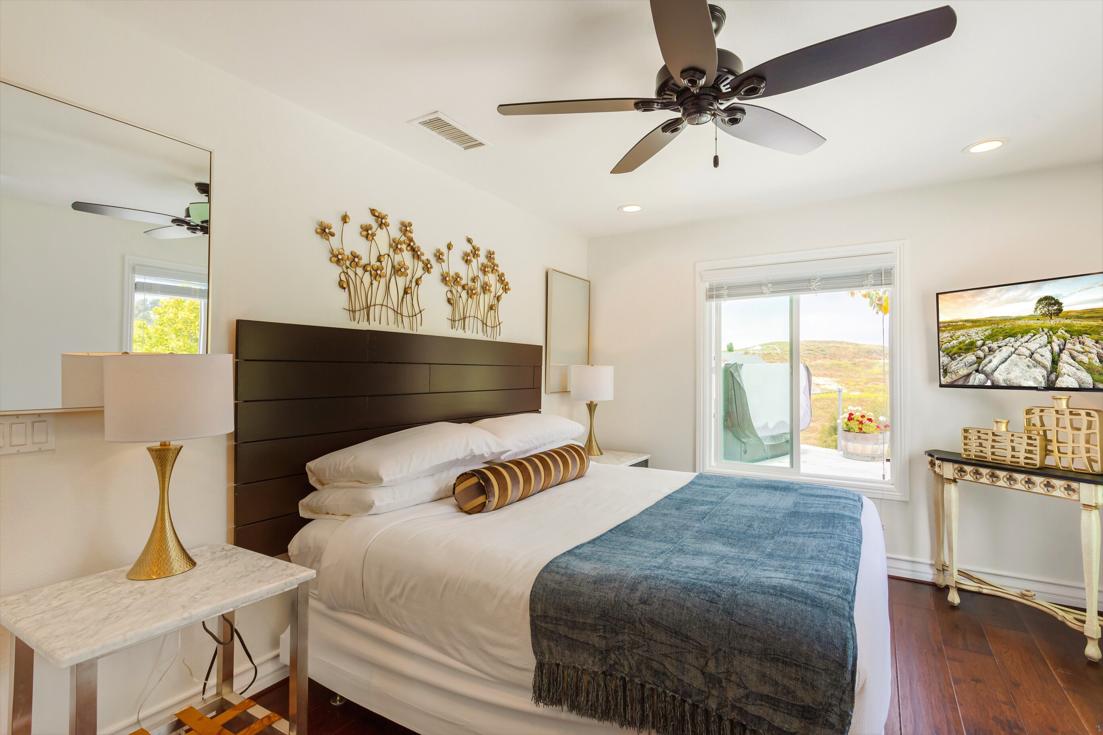 A modern bedroom with a large bed, a ceiling fan, a nightstand with a lamp, a dresser, and a window with a view of the countryside.