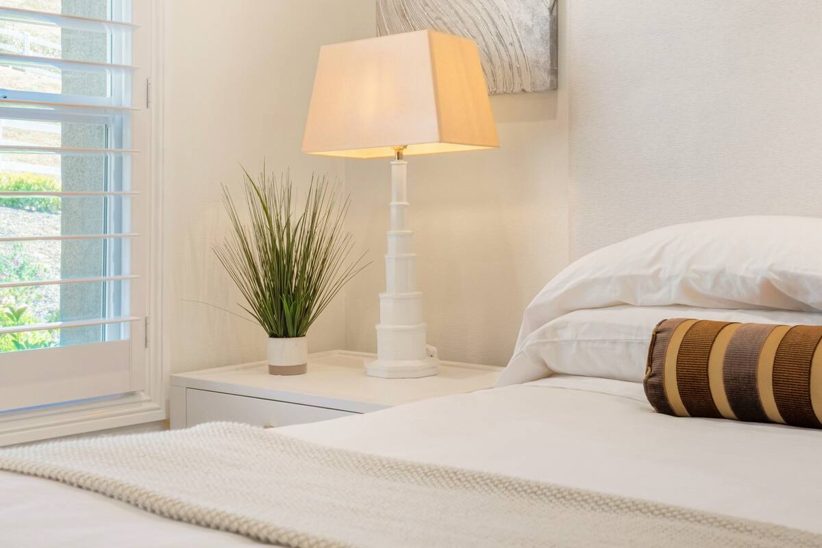 A neatly made bed with white linens and a striped pillow, a bedside lamp, and a small potted plant on a nightstand.